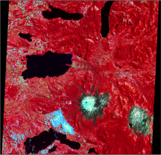 False-color satellite image showing snow-capped volcanic peaks and surrounding terrain, with vegetation in red and water bodies in black. The volcano is in the south-west center corner, flanked by lakes and rugged mountainous landscapes.
